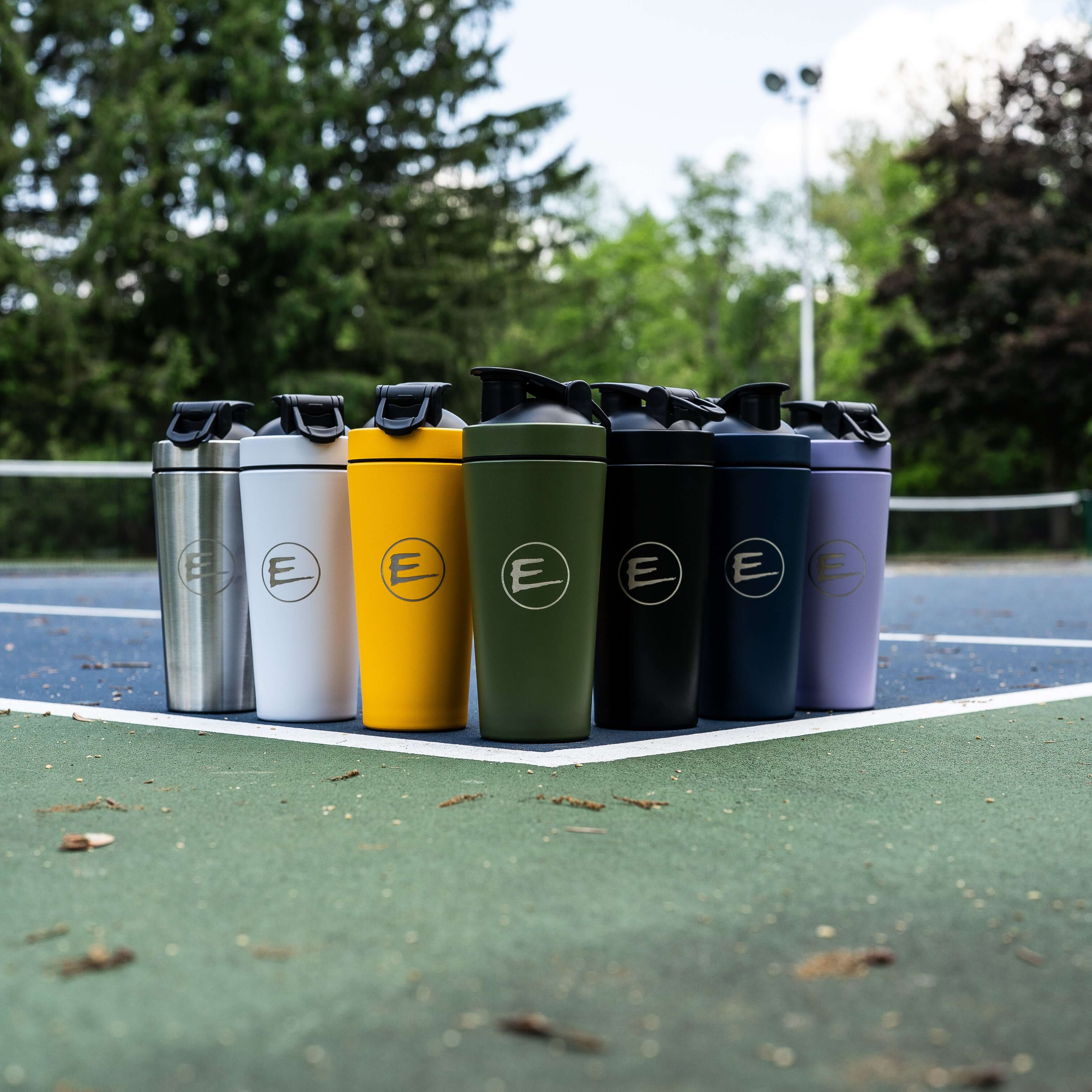 Stainless Steel 26 oz Shaker Bottle among various shaker bottles in different colors on a tennis court.